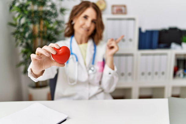 Middle age doctor woman holding heart at the clinic thinking concentrated about doubt with finger on chin and looking up wondering  - Photo, image