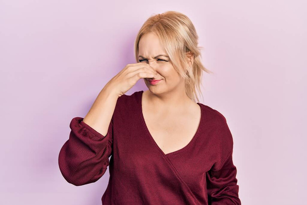 Νεαρή ξανθιά γυναίκα που φοράει ένα απλό χειμωνιάτικο πουλόβερ μυρίζοντας κάτι βρωμερό και αηδιαστικό, ανυπόφορη μυρωδιά, κρατώντας ανάσα με δάχτυλα στη μύτη. άσχημη μυρωδιά  - Φωτογραφία, εικόνα