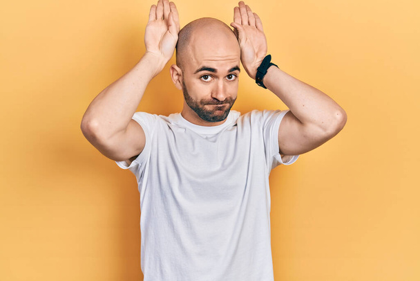Young bald man wearing casual white t shirt doing bunny ears gesture with hands palms looking cynical and skeptical. easter rabbit concept.  - Photo, Image