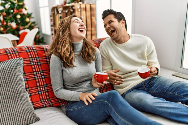 Молодая латиноамериканская пара, улыбающаяся счастливая, пьющая кофе, сидя дома на диване.. - Фото, изображение
