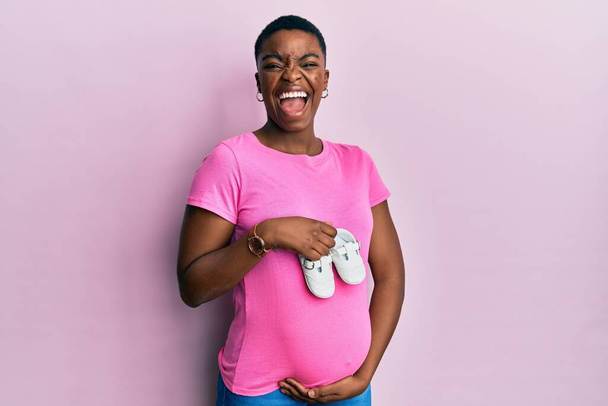 Young african american woman expecting a baby holding shoes celebrating crazy and amazed for success with open eyes screaming excited.  - Foto, imagen