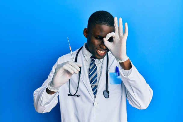 Молодой афроамериканец, доктор, держит шприц, улыбается счастливо, делает хорошо знак с рукой на глазу глядя сквозь пальцы  - Фото, изображение