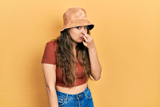 Jeune fille hispanique portant des vêtements décontractés et un chapeau sentant quelque chose de puant et dégoûtant, odeur intolérable, retenant souffle avec les doigts sur le nez. mauvaise odeur  - Photo, image