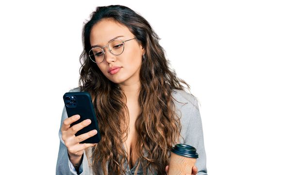 Young hispanic girl using smartphone and drinking a cup of coffee relaxed with serious expression on face. simple and natural looking at the camera.  - Photo, Image