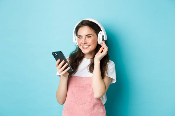 Élégante fille brune dans les écouteurs tenant smartphone, écouter de la musique, piste de sélection dans l'application de streaming, debout sur fond bleu - Photo, image