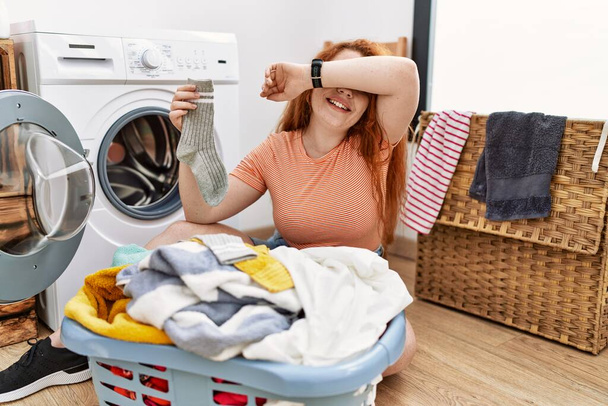 Mulher ruiva jovem colocando roupa suja na máquina de lavar roupa sorrindo alegre jogando espreitar um boo com as mãos mostrando rosto. surpreso e saiu  - Foto, Imagem