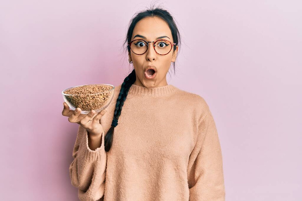 Young hispanic girl holding lentils bowl scared and amazed with open mouth for surprise, disbelief face  - Φωτογραφία, εικόνα