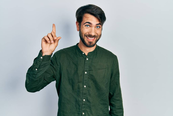 Jonge Latijns-Amerikaanse man in casual kleding glimlachend met een idee of vraag wijzend vinger omhoog met gelukkig gezicht, nummer een  - Foto, afbeelding