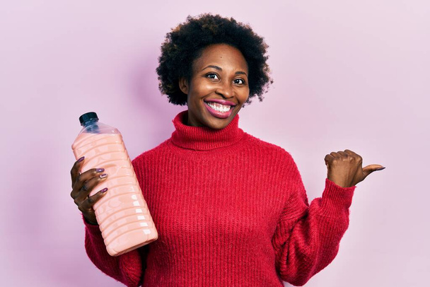 Junge afrikanisch-amerikanische Frau hält Spülmittelflasche in der Hand und zeigt Daumen zur Seite, lächelt glücklich mit offenem Mund  - Foto, Bild