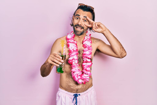 Jonge Spaanse man in badpak en Hawaïaanse lei drinken tropische cocktail doen vrede symbool met vingers over het gezicht, glimlachend vrolijk tonen overwinning  - Foto, afbeelding