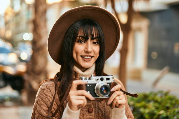 Brunette femme portant chapeau d'hiver souriant à l'aide d'une caméra vintage en plein air à la ville - Photo, image