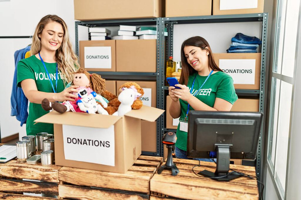 Двое молодых волонтеров улыбаются счастливо работая в благотворительном центре. Девушка улыбается счастливо с помощью смартфона. - Фото, изображение