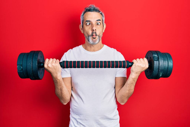 Handsome middle age man with grey hair wearing sportswear using dumbbells making fish face with mouth and squinting eyes, crazy and comical.  - Фото, изображение