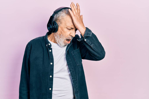 Handsome senior man with beard listening to music using headphones surprised with hand on head for mistake, remember error. forgot, bad memory concept.  - Foto, Imagem