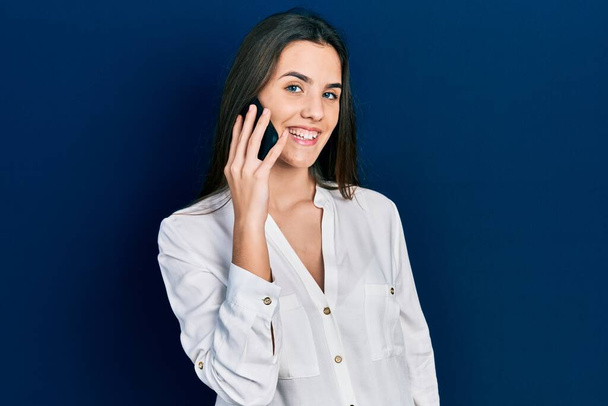 Young brunette teenager having conversation talking on the smartphone looking positive and happy standing and smiling with a confident smile showing teeth  - Photo, image