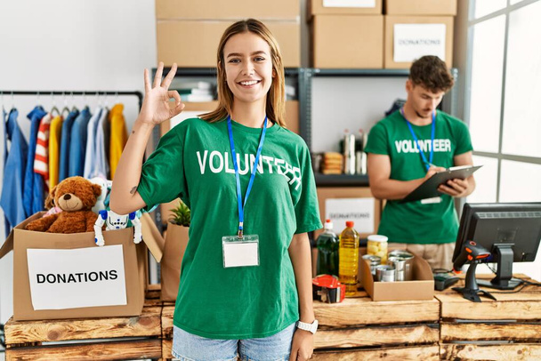 Młoda blondynka nosząca koszulkę wolontariusza na stoisku z datkami uśmiecha się pozytywnie robiąc znak ok dłońmi i palcami. udana ekspresja.  - Zdjęcie, obraz