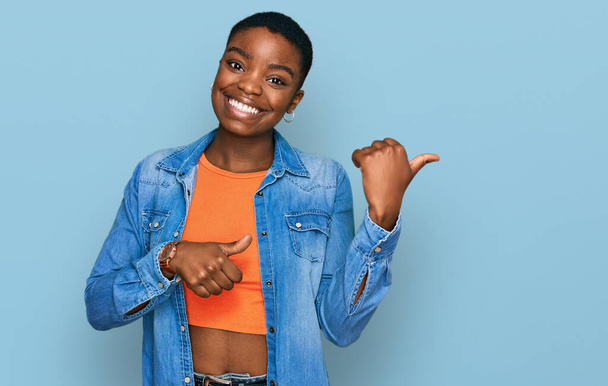 Mladá Afroameričanka v neformálním oblečení ukazuje na záda s rukou a palci nahoru, s úsměvem sebevědomá  - Fotografie, Obrázek