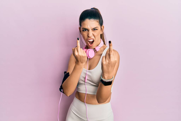 Young hispanic woman wearing gym clothes and using headphones showing middle finger doing fuck you bad expression, provocation and rude attitude. screaming excited  - Fotó, kép