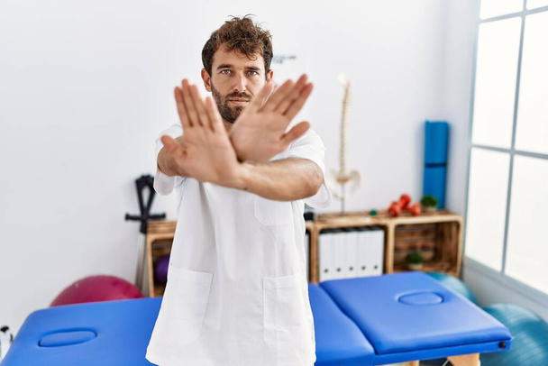 Young handsome physiotherapist man working at pain recovery clinic rejection expression crossing arms and palms doing negative sign, angry face  - 写真・画像