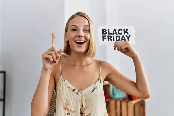 Jonge blanke vrouw met zwarte vrijdag spandoek in de winkel glimlachend met een idee of vraag wijzend met een vrolijk gezicht, nummer een  - Foto, afbeelding