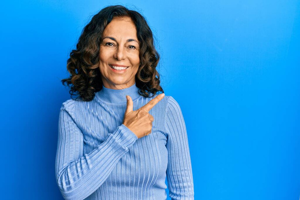Латиноамериканка средних лет в непринужденной одежде с улыбкой на лице, указывающей рукой и пальцем в сторону, со счастливым и естественным выражением лица  - Фото, изображение