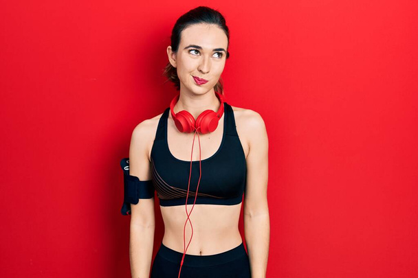 Νεαρή Ισπανίδα που φοράει ρούχα γυμναστικής και φοράει ακουστικά χαμογελώντας κοιτώντας στο πλάι και κοιτάζοντας αλλού σκεπτόμενη.  - Φωτογραφία, εικόνα