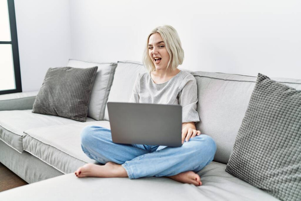 młoda kaukaska kobieta za pomocą laptopa w domu siedzi na kanapie mrugając patrząc w aparat z sexy ekspresja, wesoły i szczęśliwy twarz.  - Zdjęcie, obraz