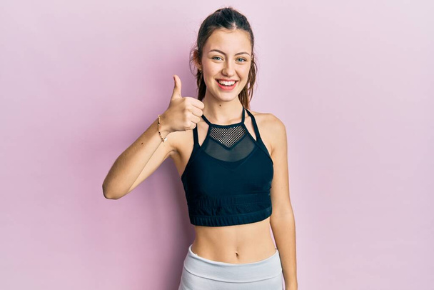 Młoda brunetka kobieta w odzieży sportowej uśmiechnięta szczęśliwy i pozytywny, kciuk do góry robi doskonałe i znak aprobaty  - Zdjęcie, obraz