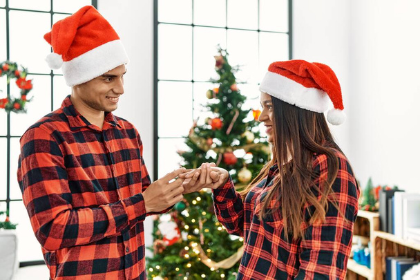 Молодая латинская пара празднует Рождество держа обручальное кольцо для свадебного предложения дома. - Фото, изображение