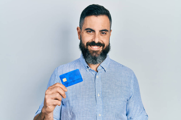 Junger hispanischer Mann mit Kreditkarte sieht im Stehen positiv und glücklich aus und lächelt mit einem selbstbewussten Lächeln, das Zähne zeigt  - Foto, Bild