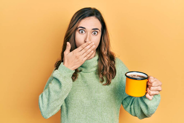 Jeune fille hispanique boire une tasse de café couvrant la bouche avec la main, choquée et effrayée par l'erreur. expression surprise  - Photo, image