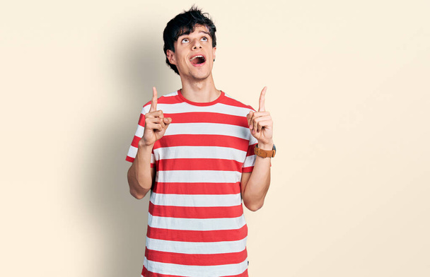 Handsome hipster young man wearing casual striped t shirt amazed and surprised looking up and pointing with fingers and raised arms.  - Photo, Image