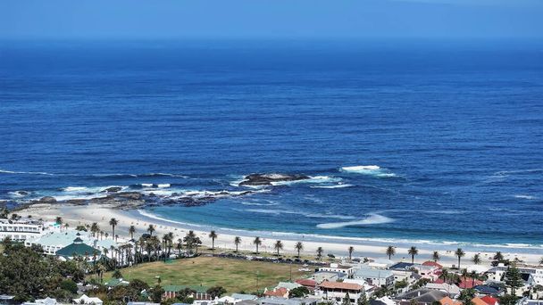 Palmeras crecen a lo largo de la playa de arena, casas de la ciudad son visibles. Las olas del azul del Océano Atlántico están espumando en la orilla. Ciudad del Cabo. Sudafrica - Foto, Imagen