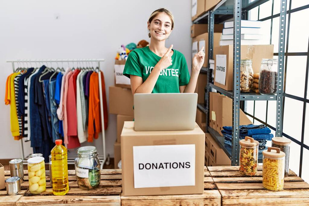 Junge blonde Frau im freiwilligen T-Shirt am Spendenstand lächelt und blickt mit zwei Händen und Fingern zur Seite in die Kamera.  - Foto, Bild