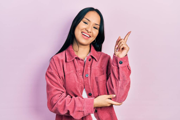 Schöne hispanische Frau mit Nasenpiercing in lässiger pinkfarbener Jacke mit einem breiten Lächeln im Gesicht, die mit Hand und Finger zur Seite in die Kamera zeigt.  - Foto, Bild