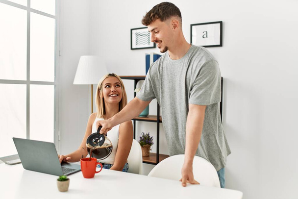 Kadın evde dizüstü bilgisayar kullanıyor ve erkek arkadaşı kahve dolduruyor.. - Fotoğraf, Görsel