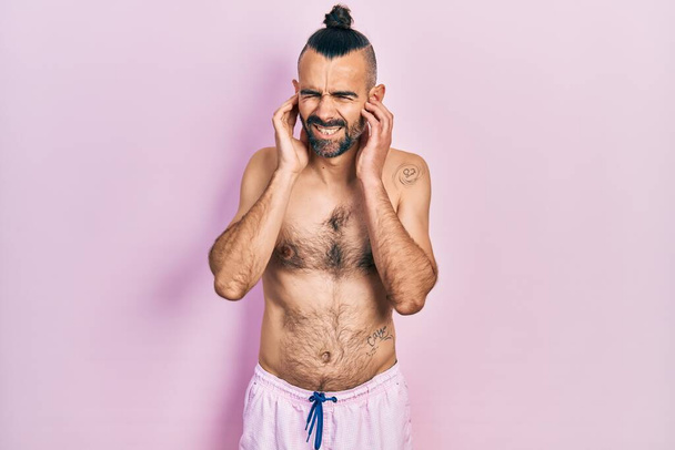 Jeune homme hispanique torse nu portant un maillot de bain couvrant les oreilles avec des doigts avec une expression agacée pour le bruit de la musique forte. concept sourd.  - Photo, image