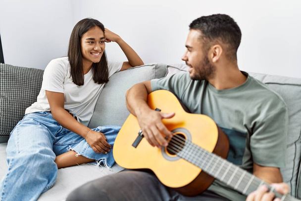 Jovem casal latino tocando guitarra clássica sentado no sofá em casa. - Foto, Imagem