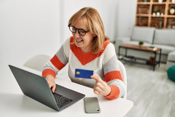 Mujer rubia de mediana edad comprando usando laptop y tarjeta de crédito en casa. - Foto, imagen