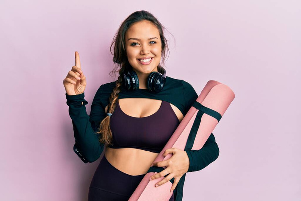 Young hispanic girl wearing sportswear holding yoga mat smiling with an idea or question pointing finger with happy face, number one  - Photo, Image