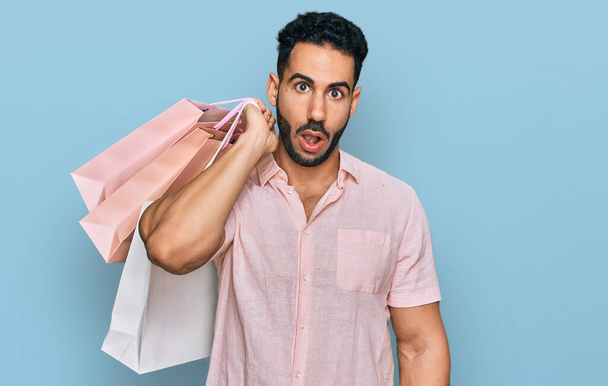 Hispanic man with beard holding shopping bags scared and amazed with open mouth for surprise, disbelief face  - Foto, imagen