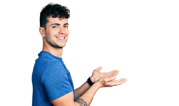 Mladý hispánec nosí ležérní tričko ukazující stranou s rukama otevřenýma dlaněmi ukazujícíma kopírovací prostor, prezentující reklamu s nadšeným úsměvem  - Fotografie, Obrázek