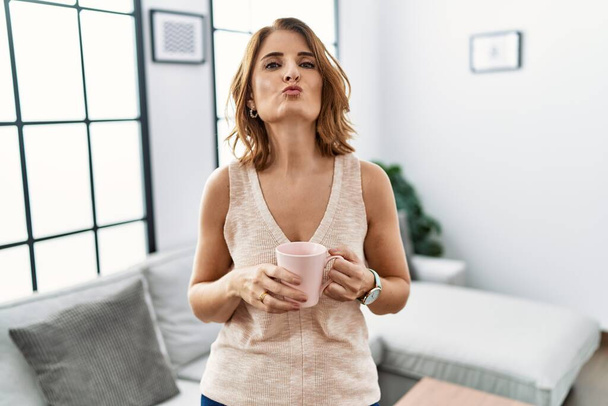 Mujer de mediana edad bebiendo una taza de café en casa mirando a la cámara soplando un beso en el aire siendo encantador y sexy. expresión de amor.  - Foto, imagen