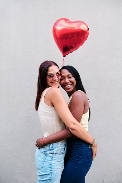 Χαρούμενο λεσβιακό ζευγάρι αγκαλιάζει κρατώντας ένα κόκκινο μπαλόνι σε σχήμα καρδιάς. - Φωτογραφία, εικόνα