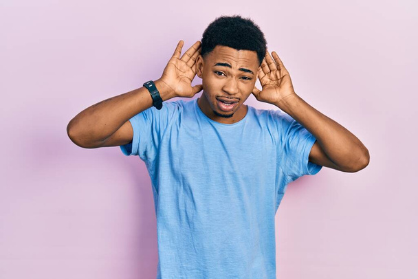 Νεαρός Αφροαμερικάνος που φοράει μπλε μπλουζάκι προσπαθώντας να ακούσει και τα δύο χέρια σε μια χειρονομία αυτιού, περίεργος για κουτσομπολιά. πρόβλημα ακοής, κωφός  - Φωτογραφία, εικόνα