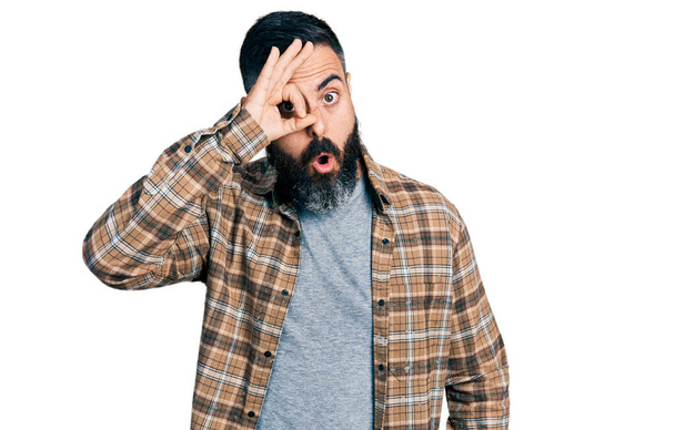 Hispanic man with beard wearing casual shirt doing ok gesture shocked with surprised face, eye looking through fingers. unbelieving expression.  - Foto, imagen