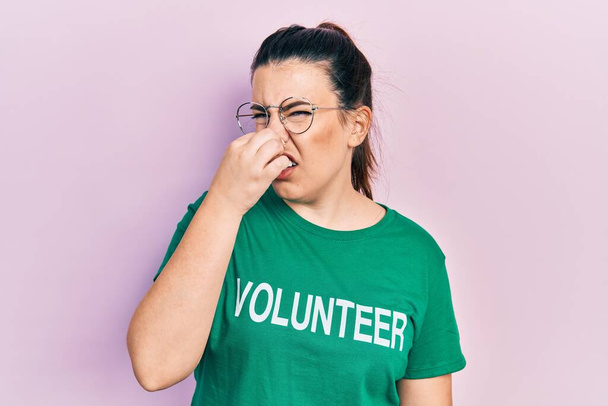 Jeune femme hispanique portant un t-shirt volontaire sentant quelque chose de puant et dégoûtant, odeur intolérable, retenant son souffle avec les doigts sur le nez. mauvaise odeur  - Photo, image