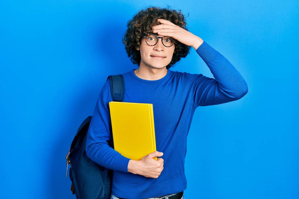 Handsome young man wearing student backpack and holding book stressed and frustrated with hand on head, surprised and angry face  - Photo, image