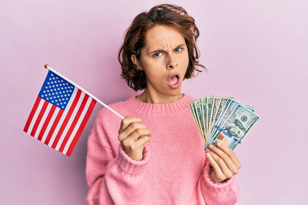 Junge brünette Frau mit US-Flagge und Dollars im Schockgesicht, skeptisch und sarkastisch guckend, überrascht mit offenem Mund  - Foto, Bild