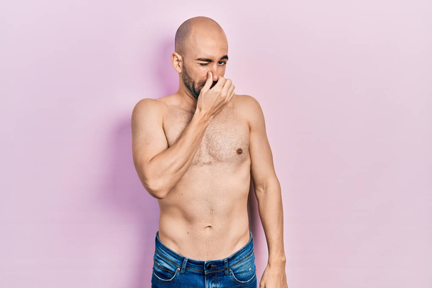 Young bald man standing shirtless smelling something stinky and disgusting, intolerable smell, holding breath with fingers on nose. bad smell  - Foto, Imagem